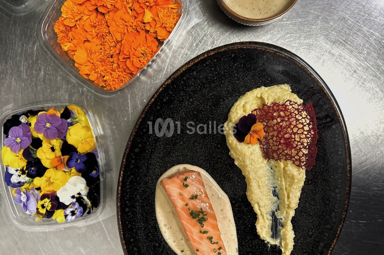 Assiette gastronomique avec saumon, purée, sauce et décoration florale, entourée de fleurs comestibles en contenants.