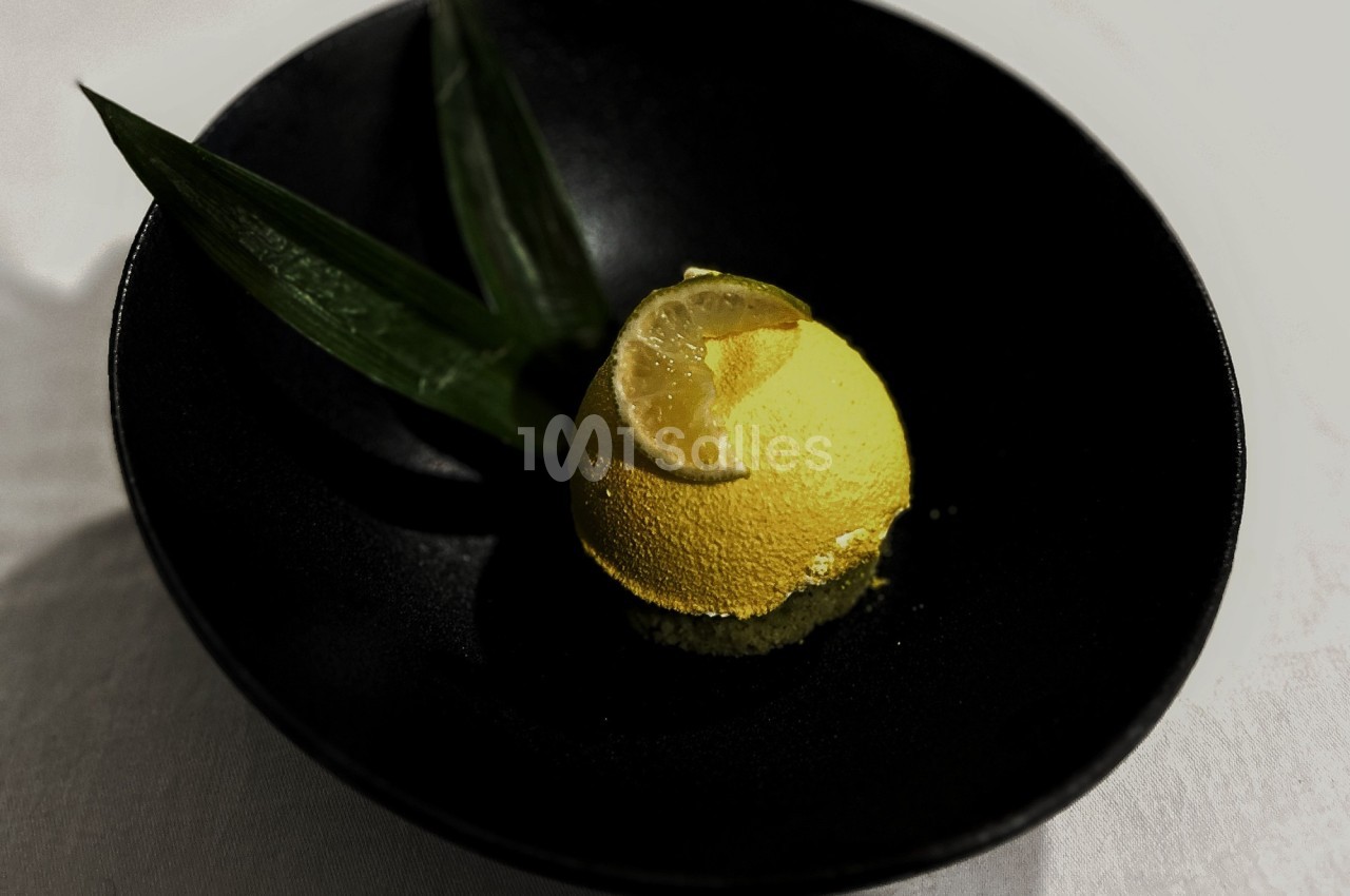 Dessert jaune en forme de dôme, garni d'une tranche de citron et accompagné de feuilles vertes, présenté dans une assiette…