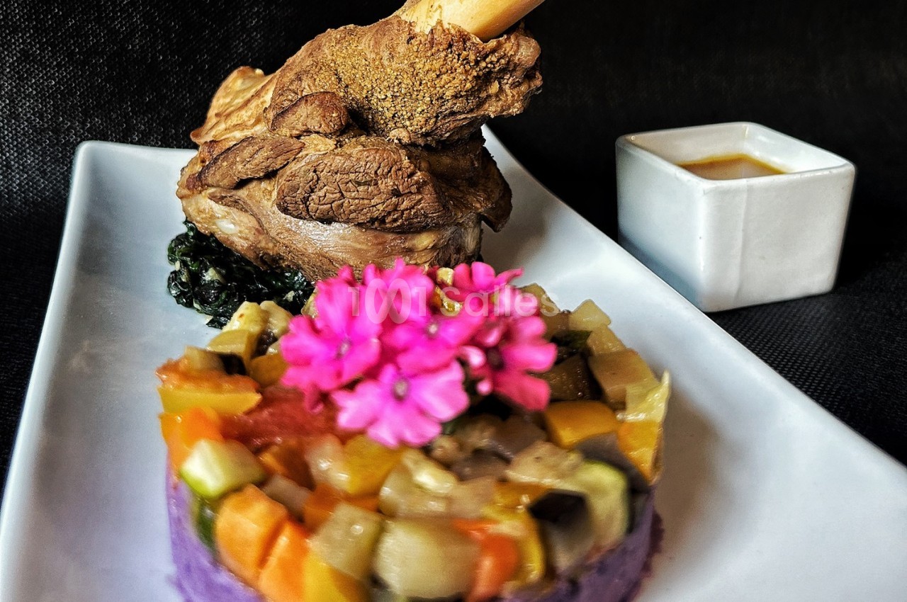Jarret d'agneau rôti sur lit de légumes colorés et purée violette, décoré de fleurs comestibles, avec une sauce à part.