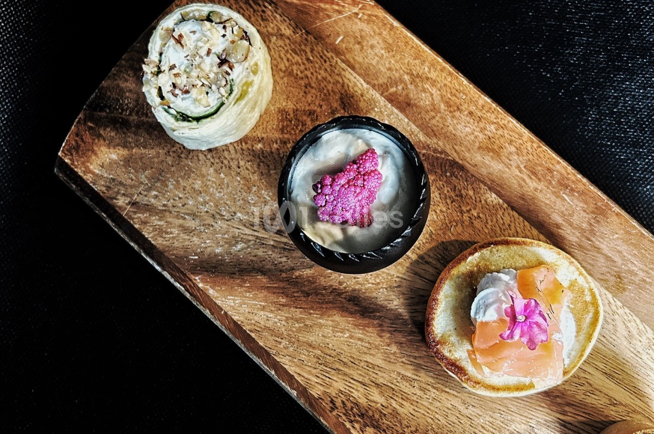 Assortiment de trois amuse-bouches disposés sur une planche en bois, comprenant saumon, mousse et garnitures variées.