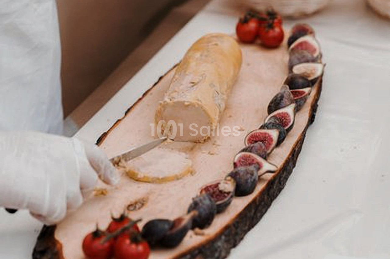 Une main coupe un rouleau de foie gras sur une planche en bois, entouré de figues et de tomates cerises.