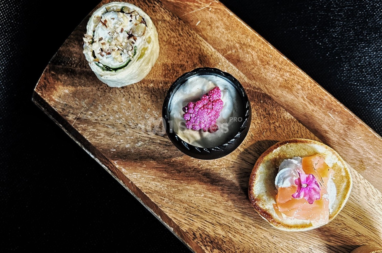 Assortiment de trois amuse-bouches disposés sur une planche en bois, comprenant saumon, mousse et garnitures variées.