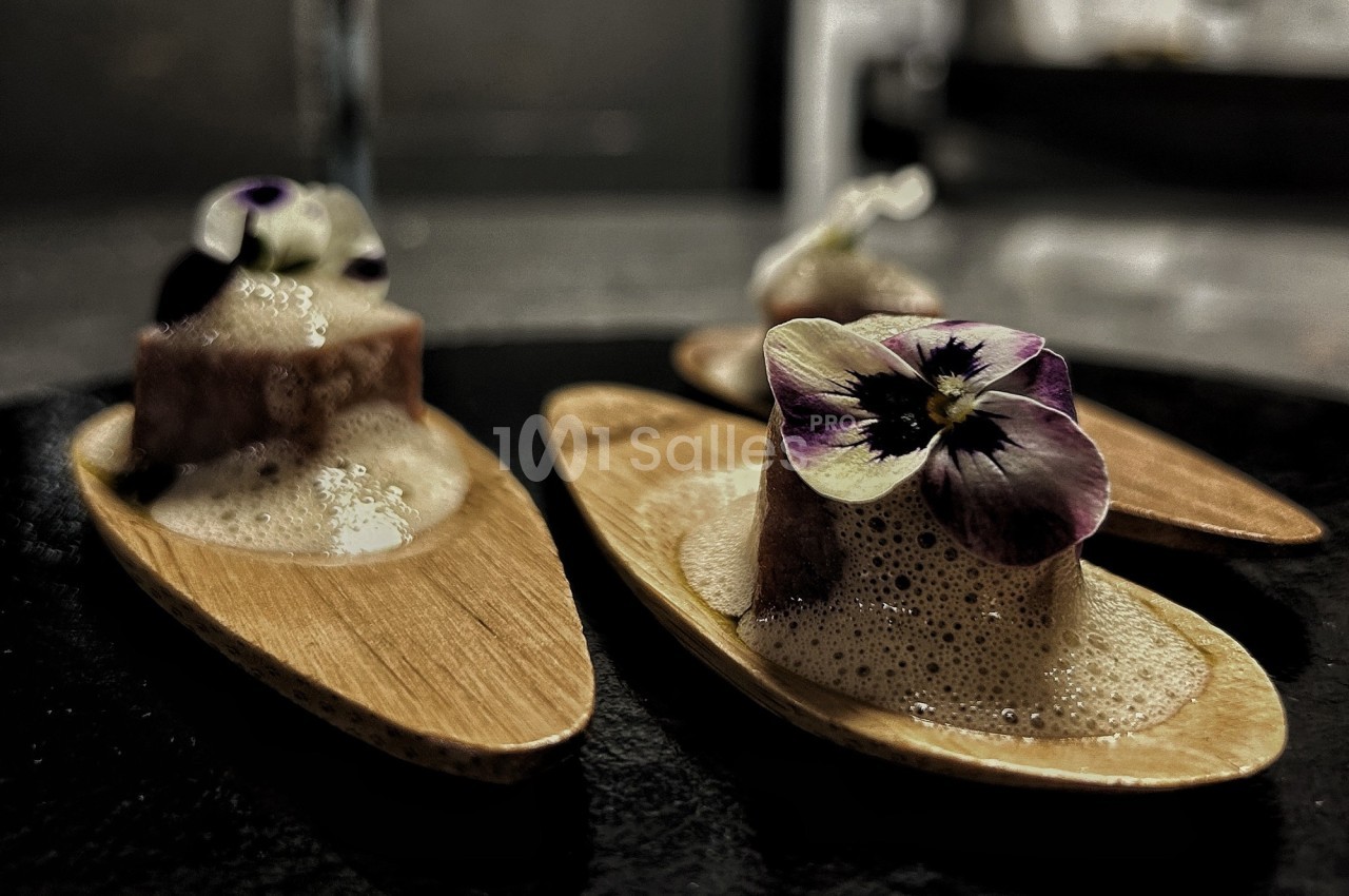 Des bouchées gastronomiques servies sur des cuillères en bois, décorées de fleurs comestibles et d'écume.