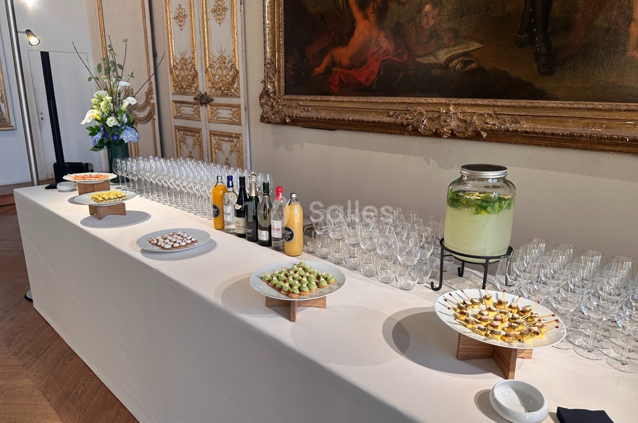 Buffet élégant avec amuse-bouches, boissons variées et décoration florale dans une salle ornée de tableaux et moulures…