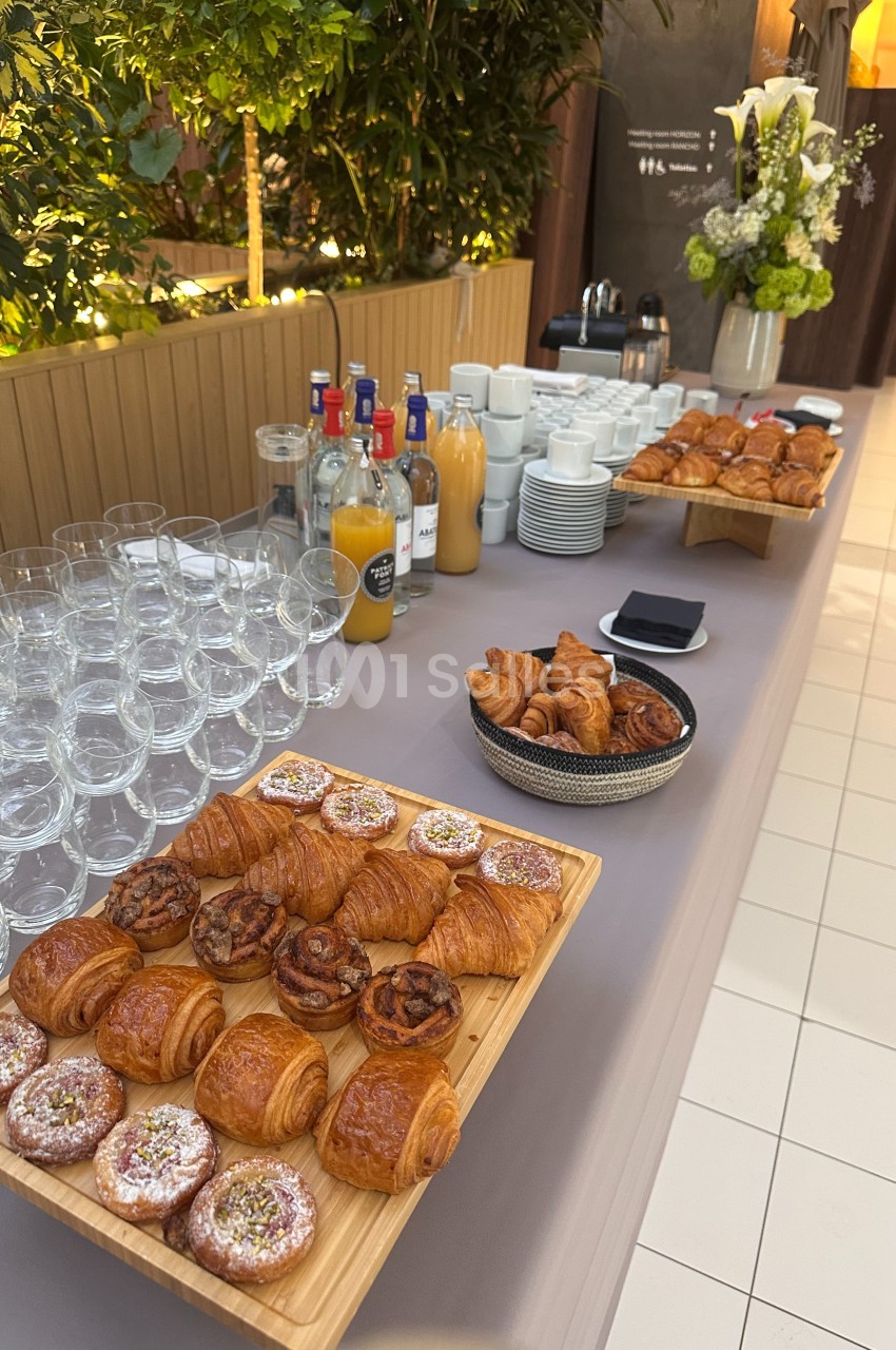 Buffet de petit-déjeuner avec viennoiseries, jus de fruits, verres et café, disposé sur une table dans un cadre lumineux.