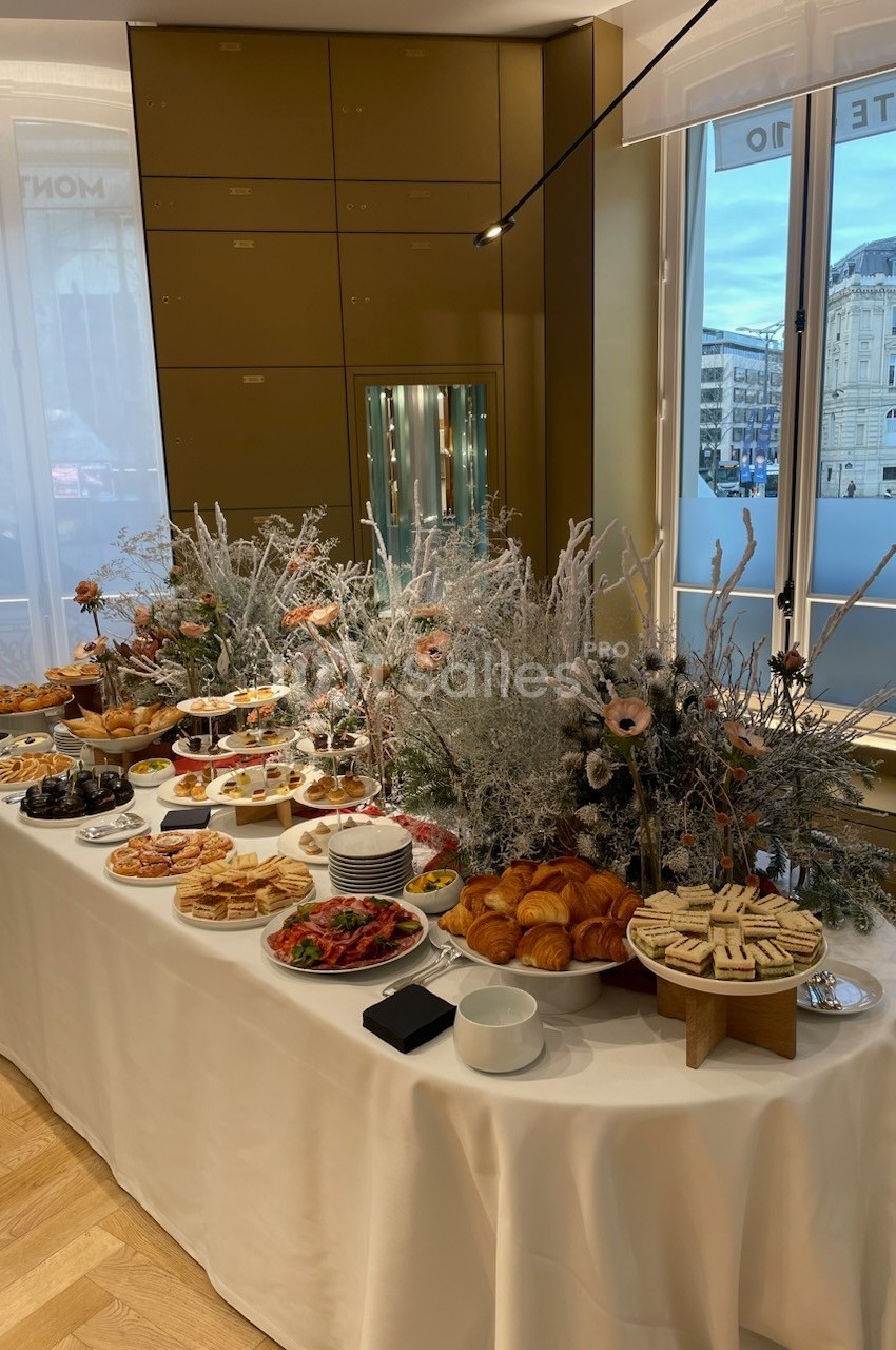 Table garnie de viennoiseries, tartes, fruits et autres mets, décorée de fleurs et branches hivernales, près de fenêtres.