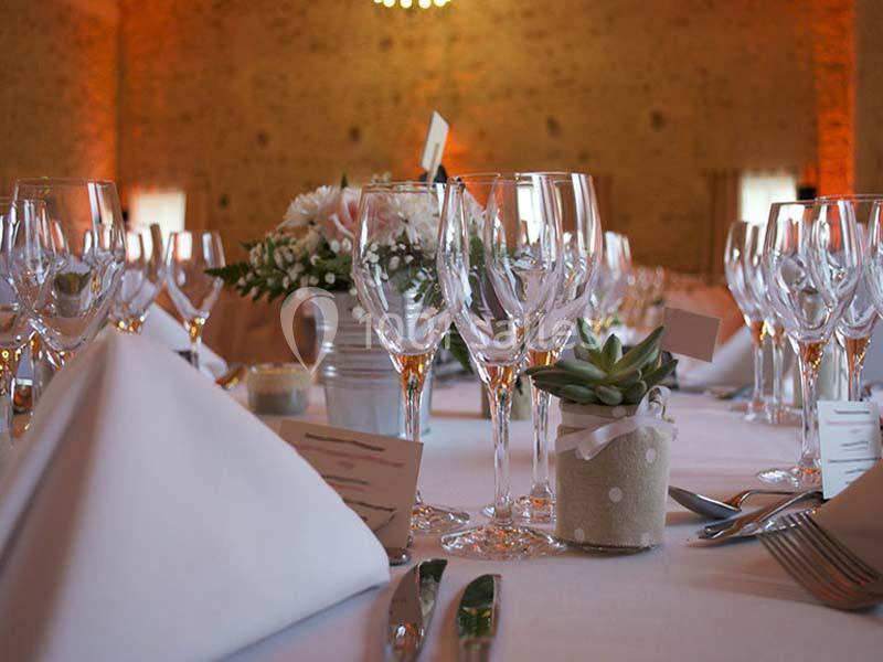 Table dressée avec nappes blanches, verres à vin, serviettes pliées, fleurs et une petite plante en pot en décoration.