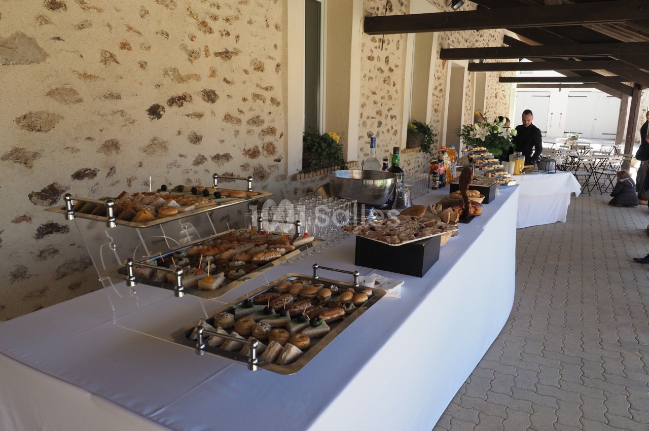Buffet en extérieur avec viennoiseries, desserts, boissons et verres disposés sur une table blanche près d'un mur en pierre.