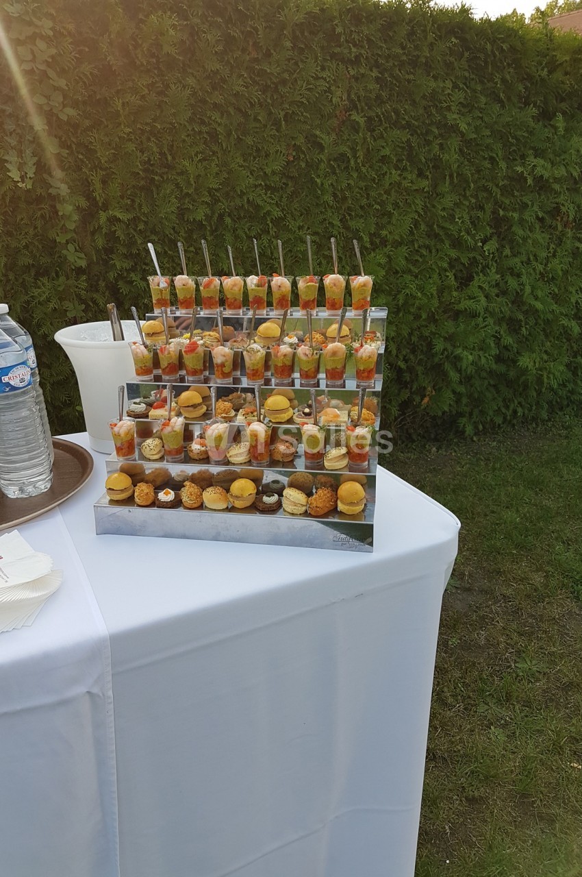 Présentoir de verrines et amuse-bouches variés sur une table blanche en extérieur, près d'une haie verte.