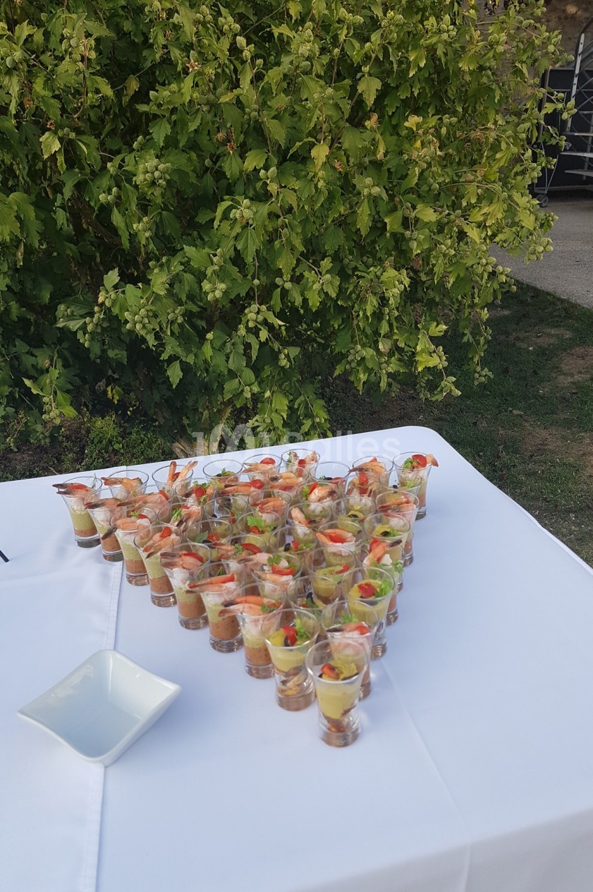 Verrines apéritives disposées en triangle sur une table blanche en extérieur, avec des arbustes en arrière-plan.