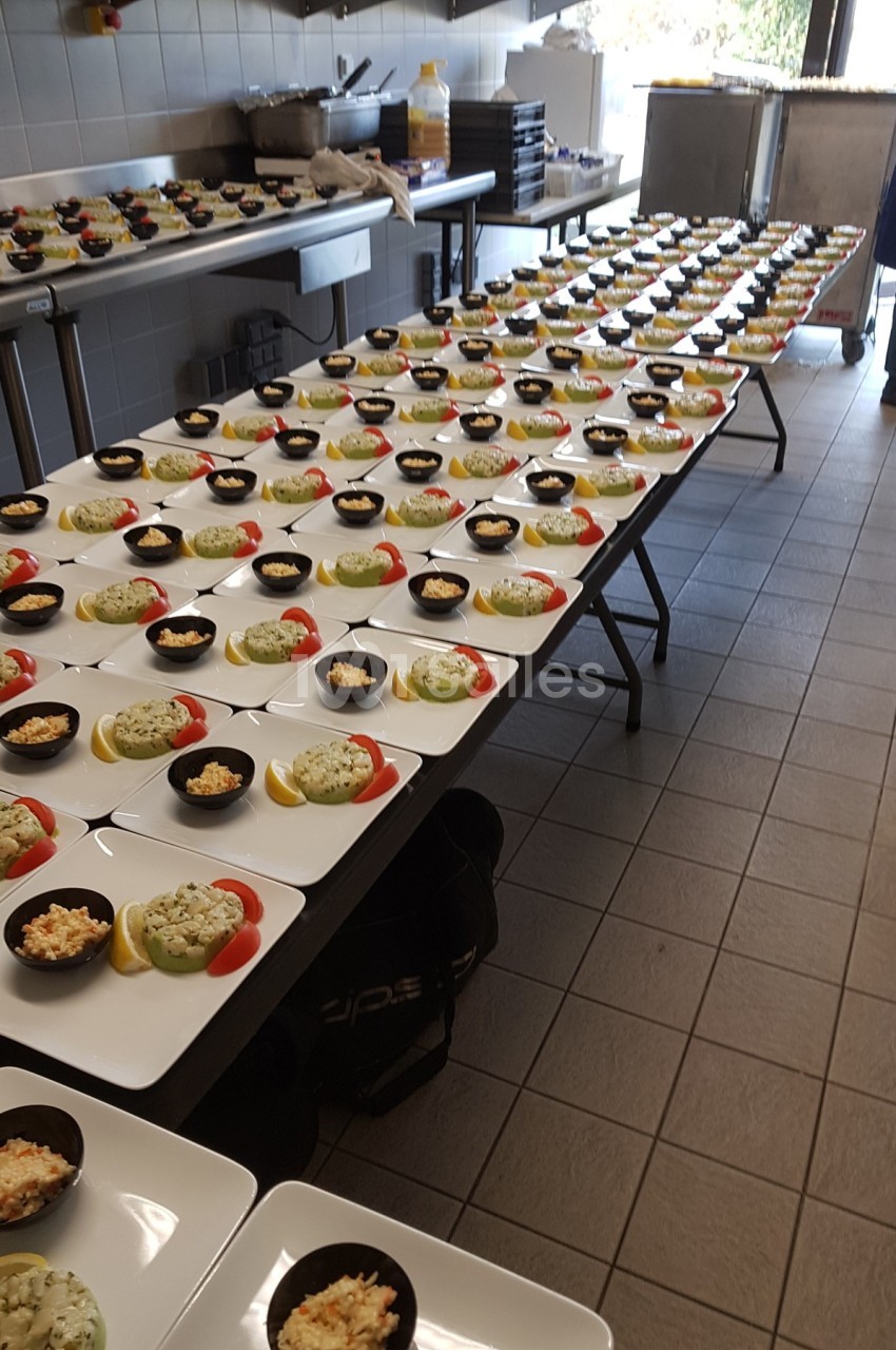 Des assiettes alignées sur des tables dans une cuisine, contenant des entrées avec des verrines et des garnitures colorées.