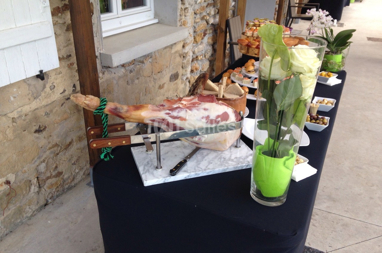 Jambon entier sur un support avec couteaux, disposé sur une table de buffet à côté de fleurs et d'amuse-bouches.