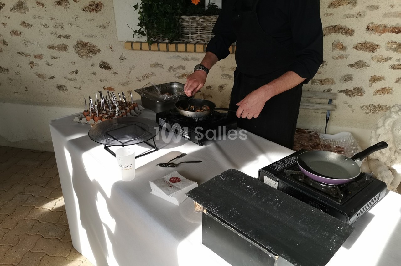 Un cuisinier prépare un plat sur une plaque chauffante lors d'un événement en extérieur, table dressée avec ustensiles.
