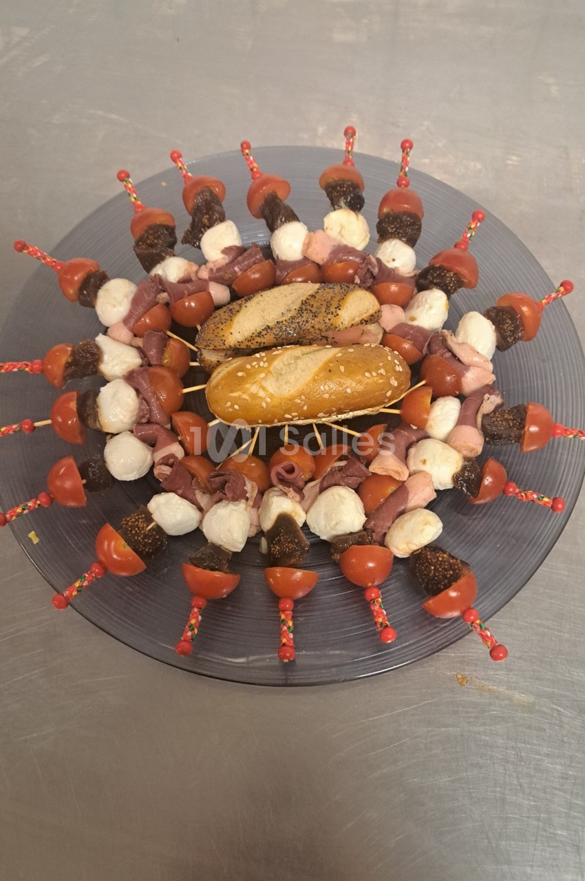 Assiette circulaire garnie de brochettes apéritives avec tomates, mozzarella, charcuterie et deux petits pains au centre.