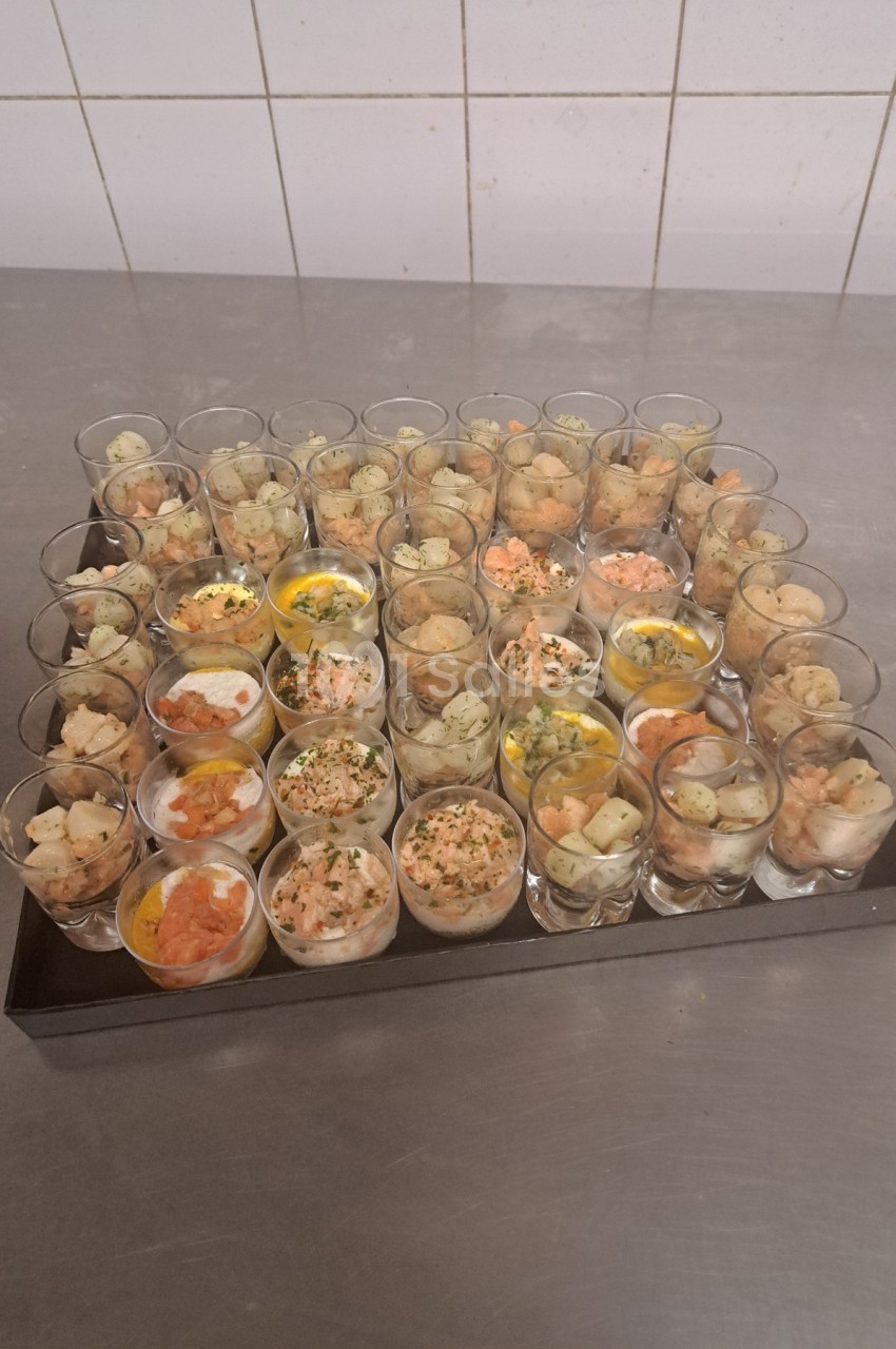 Plateau de verrines variées contenant des ingrédients comme du saumon, des œufs et des herbes, posé sur une table en inox.