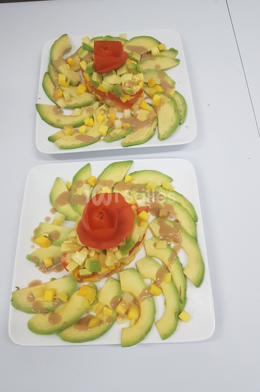 Assiette garnie de tranches d'avocat, dés de mangue et tomate sculptée en rose, présentée sur une table blanche.