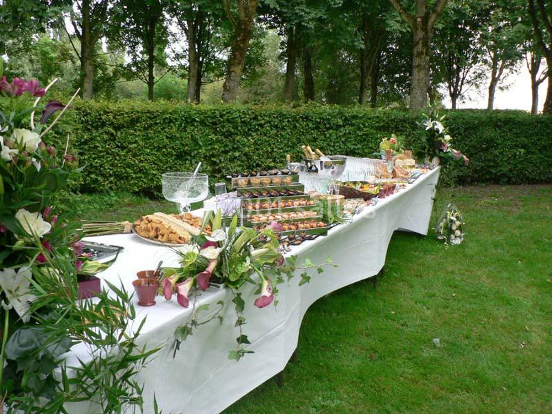 Table de buffet dressée en extérieur, garnie de plats variés, décorée de fleurs et entourée de verdure.
