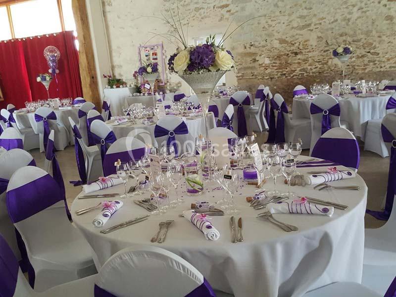Salle décorée pour un événement avec des tables rondes dressées, nappes blanches et accents violets.