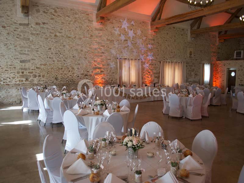 Salle de réception avec murs en pierre, tables rondes décorées et chaises blanches sous un plafond en poutres apparentes.