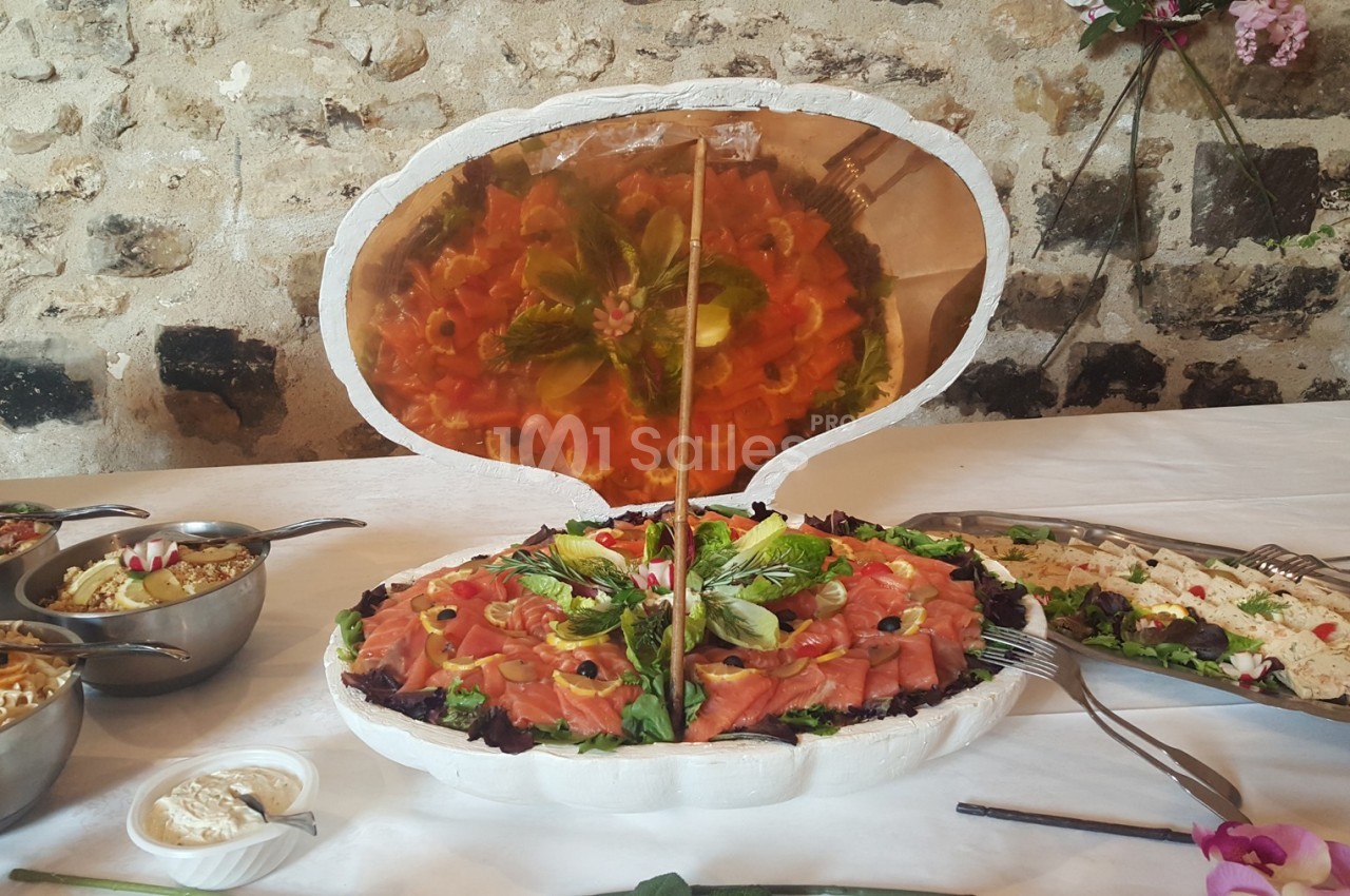 Plateau de saumon fumé présenté dans une coquille décorative, entouré de divers plats sur une table blanche.