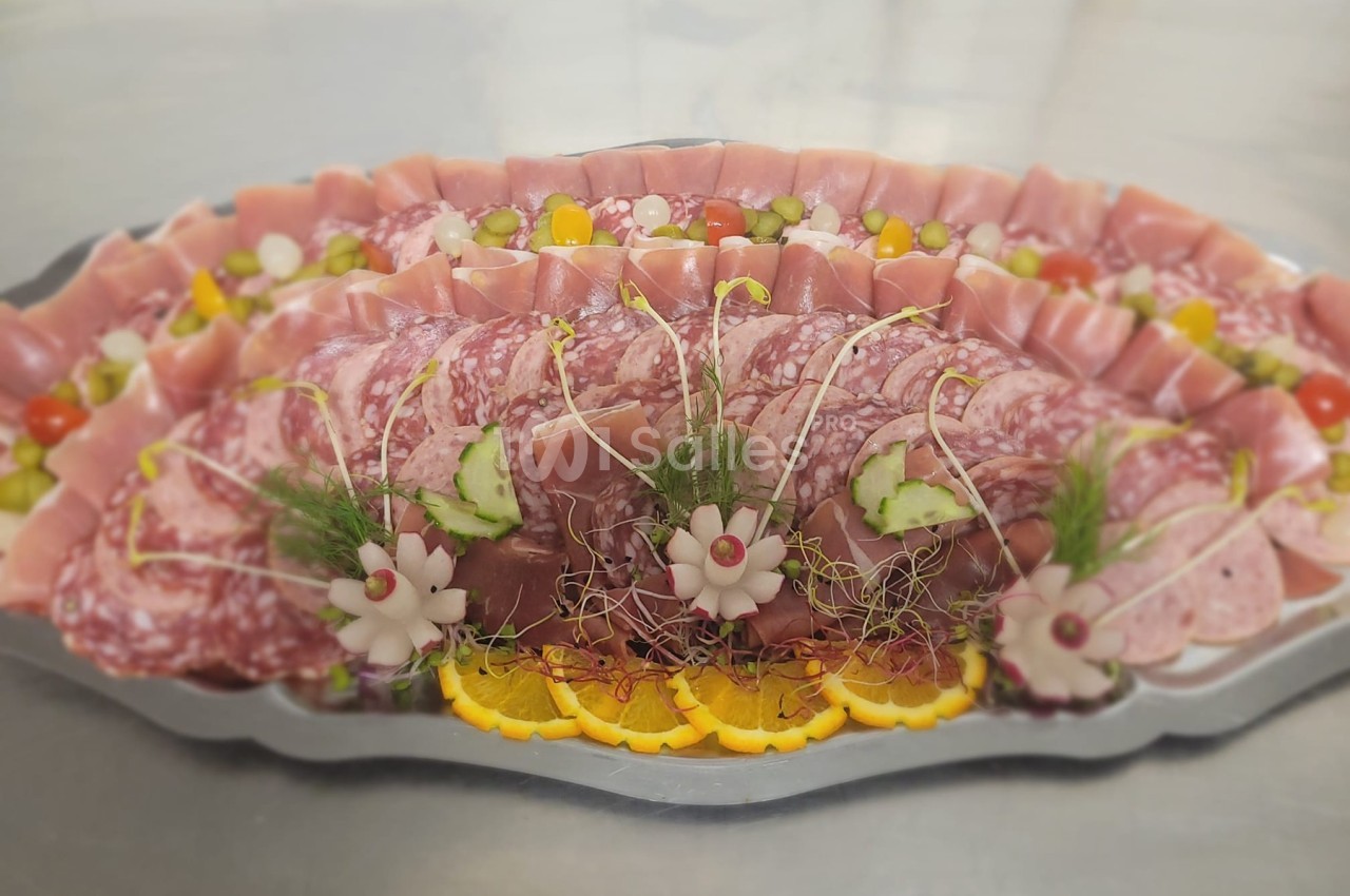 Plateau de charcuterie avec tranches de jambon, saucisson, décorations de légumes et herbes fraîches.