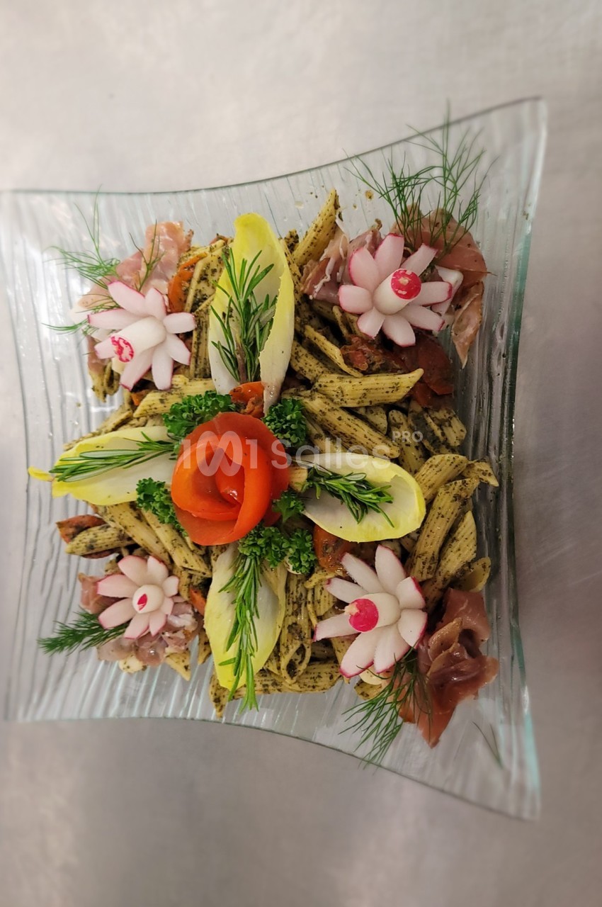Assiette de pâtes au pesto décorée de légumes sculptés, endives, herbes fraîches et tranches de jambon.