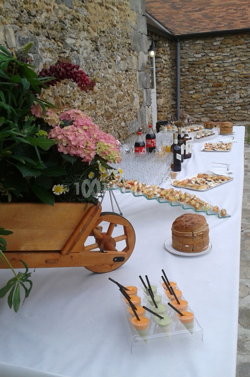 Buffet extérieur avec boissons, verrines, amuse-bouches et décor floral sur une table blanche près d'un mur en pierre.