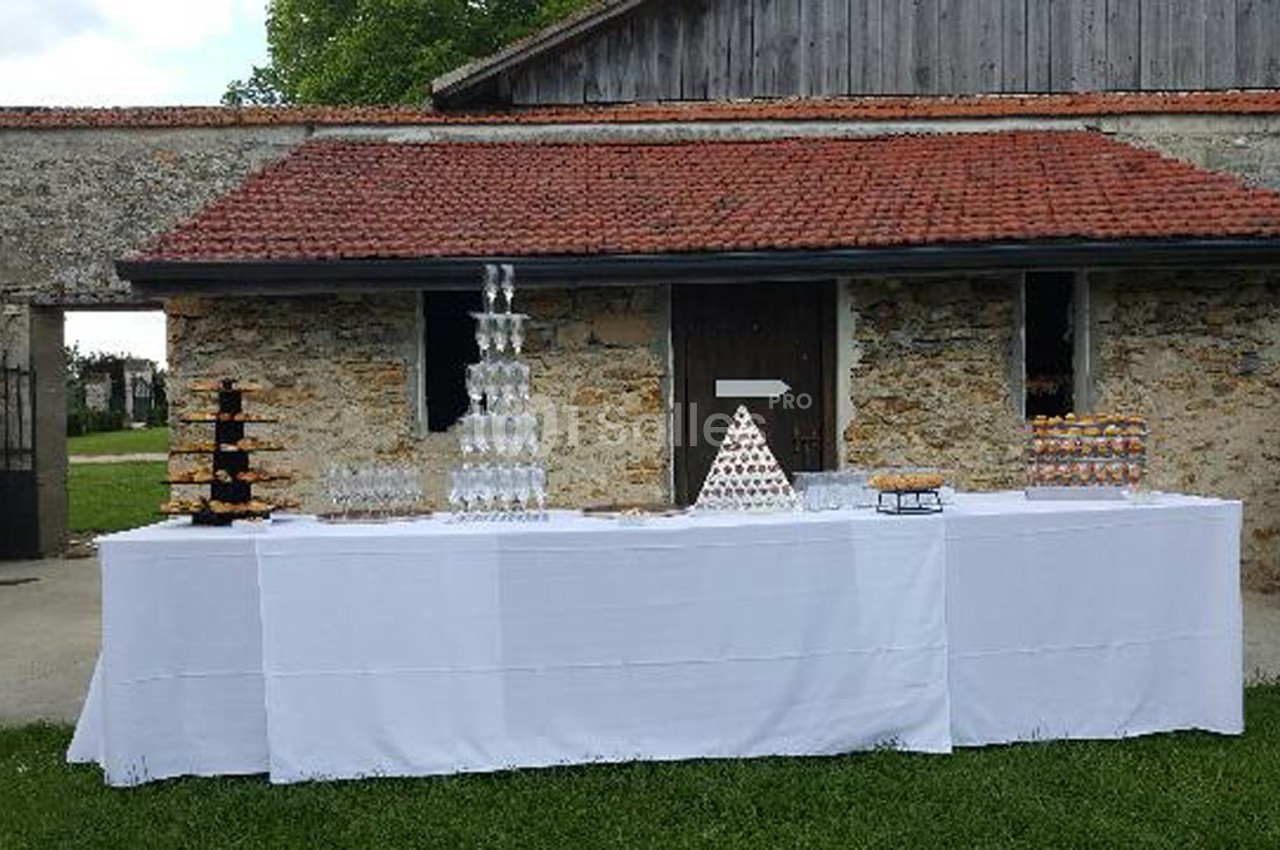 Table de réception en extérieur avec nappes blanches, pyramides de verres et de desserts devant un bâtiment en pierre.
