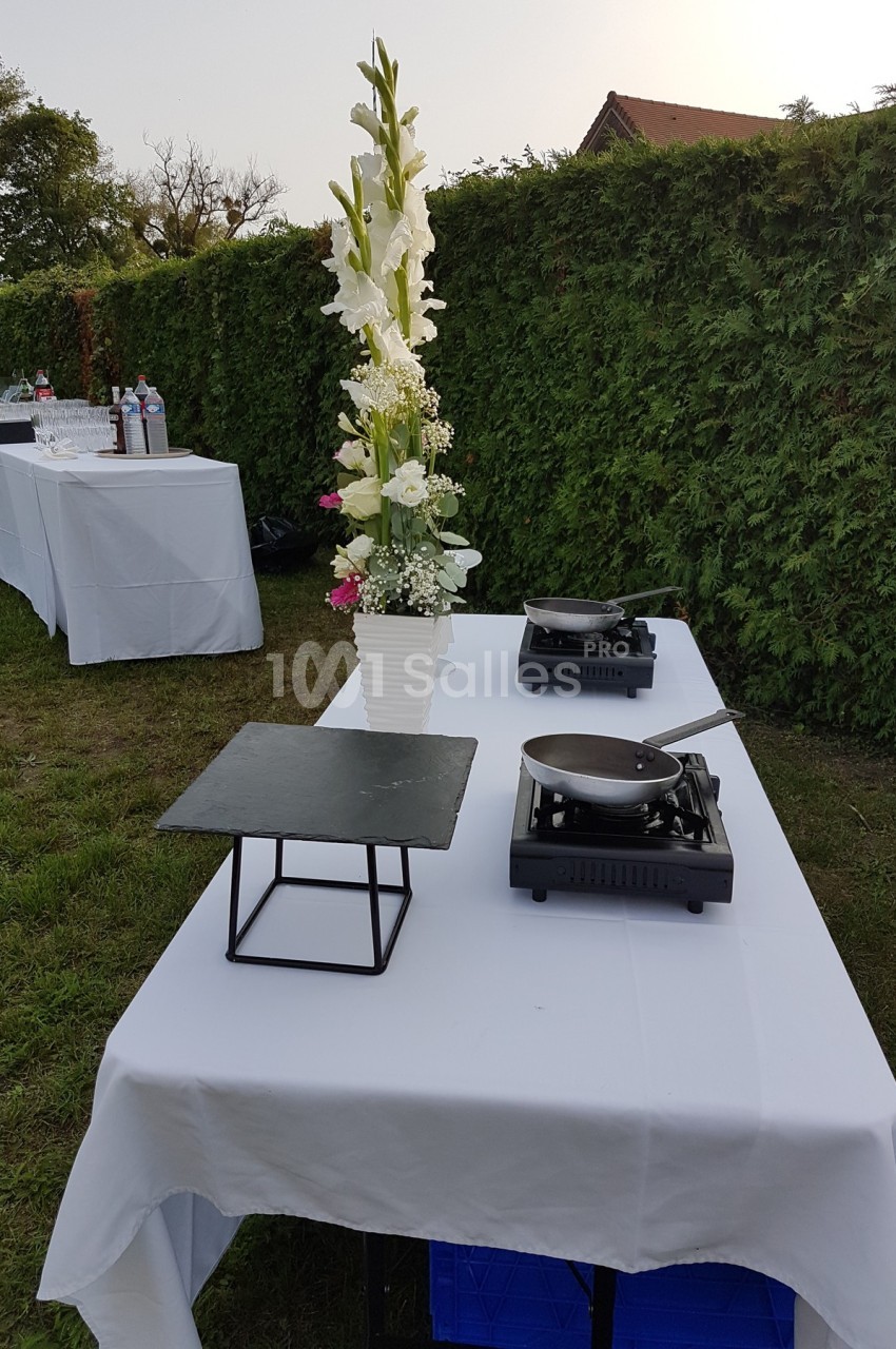 Table dressée en extérieur avec nappes blanches, plancha, poêle, ardoise et vase de fleurs blanches.