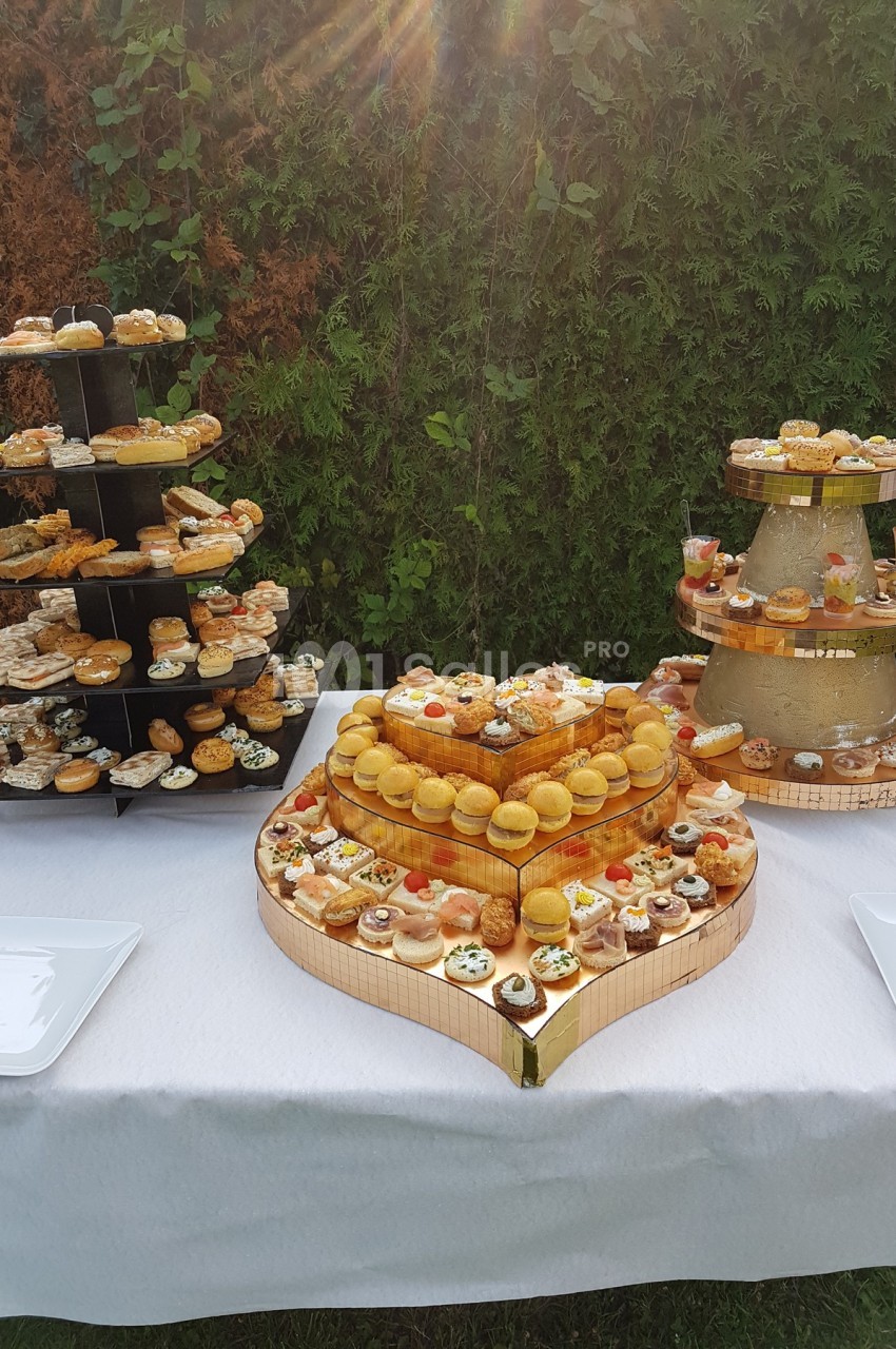 Buffet de pâtisseries et amuse-bouches disposés sur des présentoirs en extérieur, avec une nappe blanche.