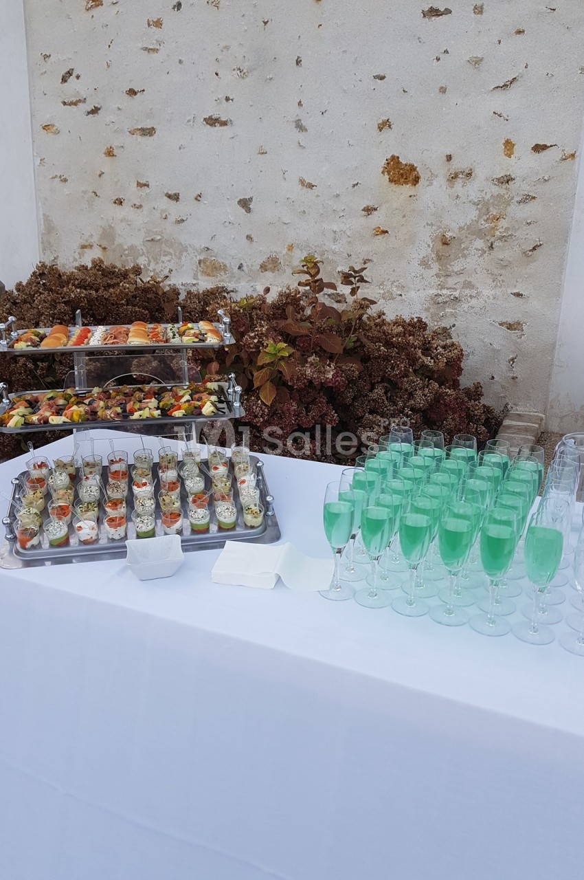 Table dressée avec verrines, plateau de fruits et verres de cocktail vert devant un mur en pierre.