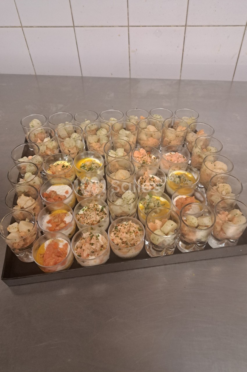 Plateau de verrines variées contenant des ingrédients comme du saumon, des œufs et des herbes, posé sur une table en inox.