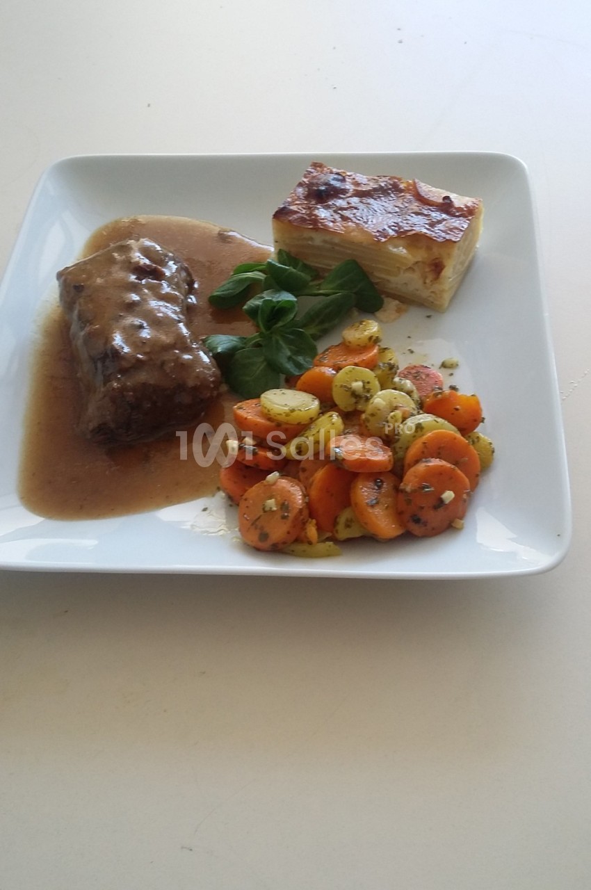 Assiette contenant un rôti en sauce, un gratin dauphinois et des légumes sautés, présentée sur une table blanche.