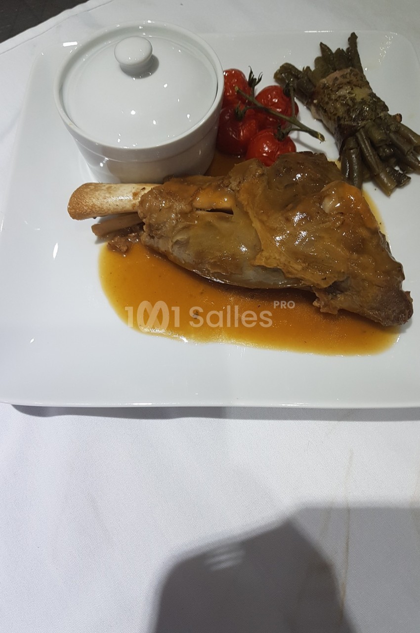 Plat avec un jarret d'agneau en sauce, tomates cerises rôties, haricots verts en fagot et une cocotte blanche.