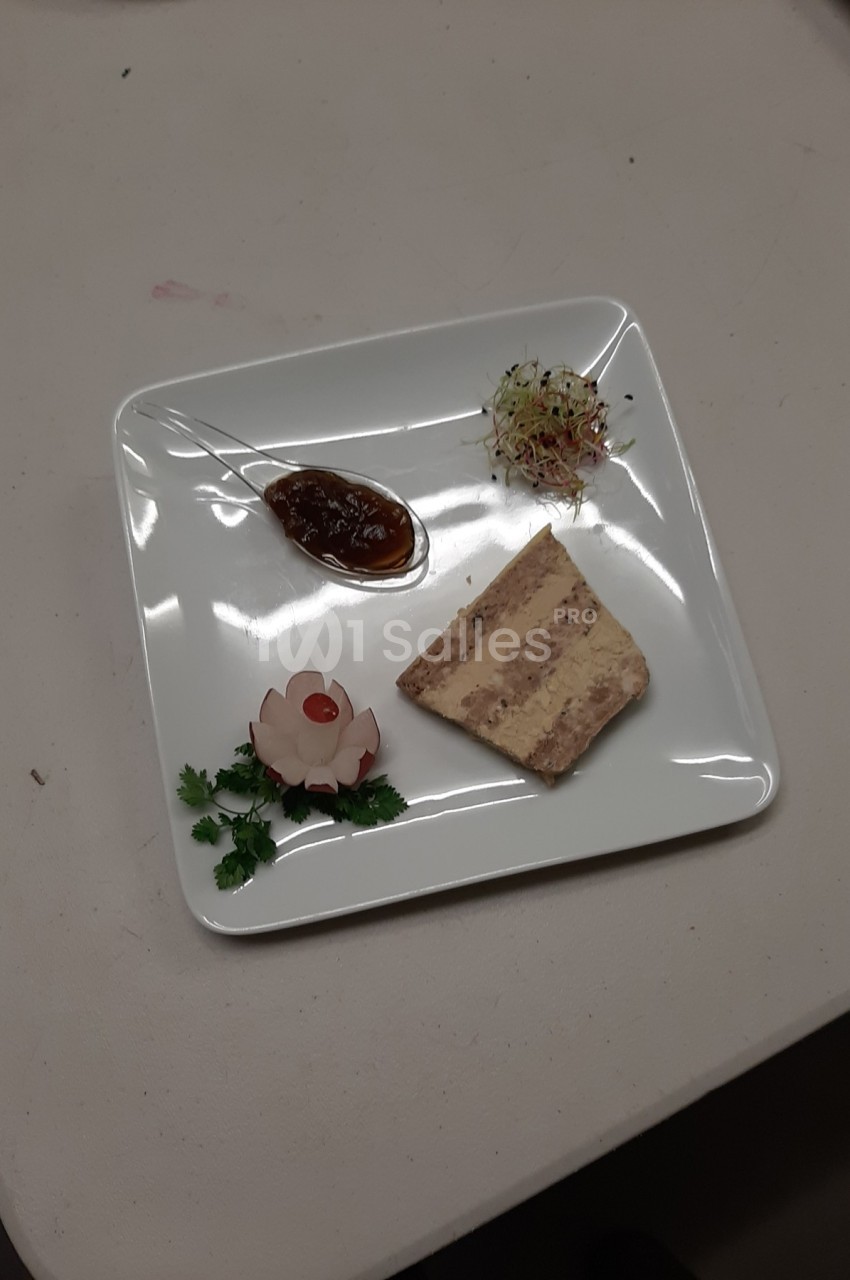 Tranche de terrine avec confiture, décoration florale en légume, herbes fraîches et germes sur une assiette blanche.