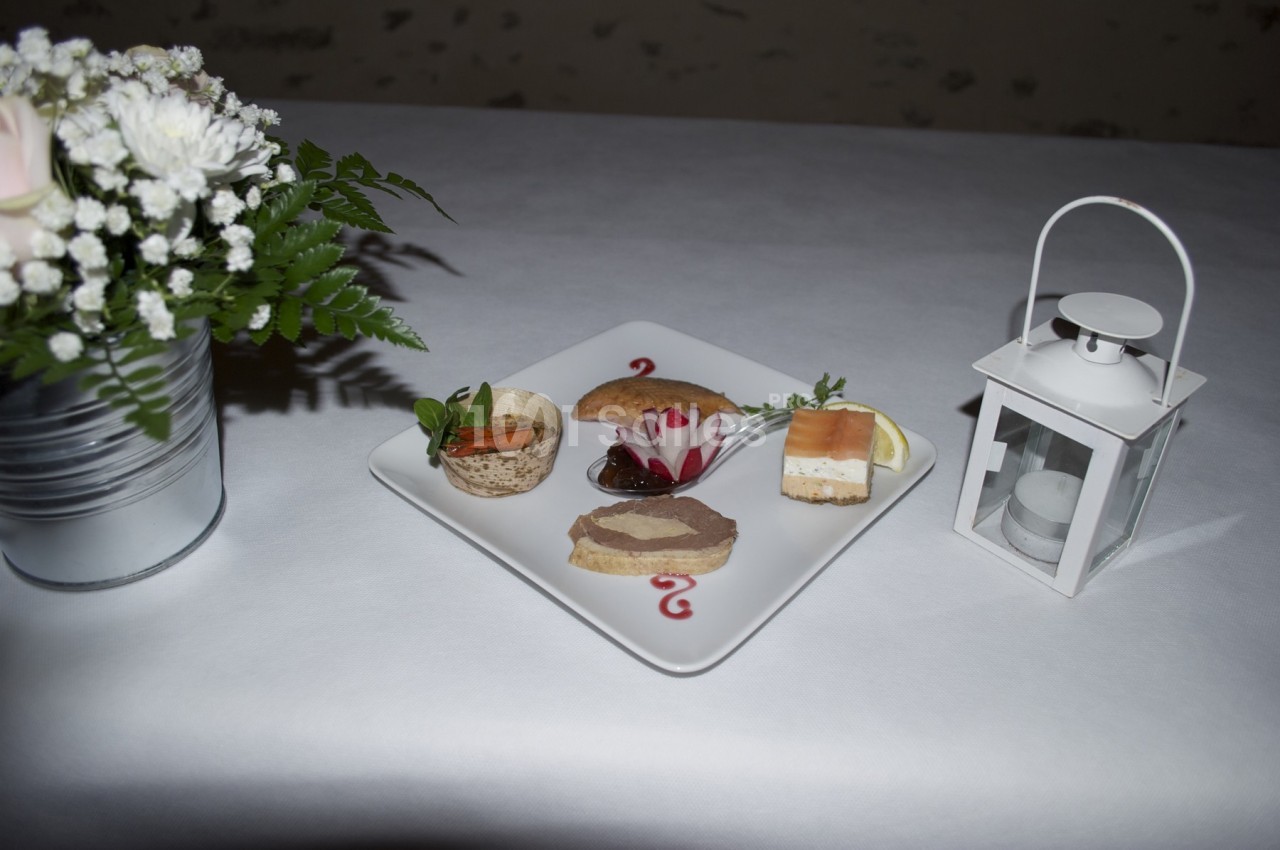 Assiette de mise en bouche variées sur une nappe blanche, avec une lanterne et un bouquet de fleurs en arrière-plan.
