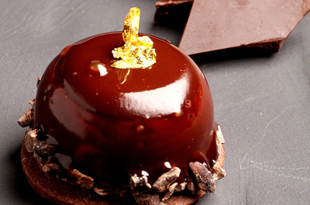 Dessert au chocolat brillant orné de feuille d'or, posé sur une base croquante avec éclats de cacao.