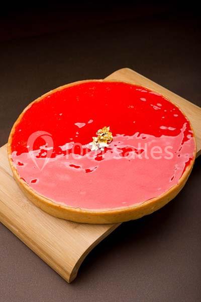 Tarte brillante au glaçage rouge vif, décorée de feuilles d'or, posée sur une planche en bois.