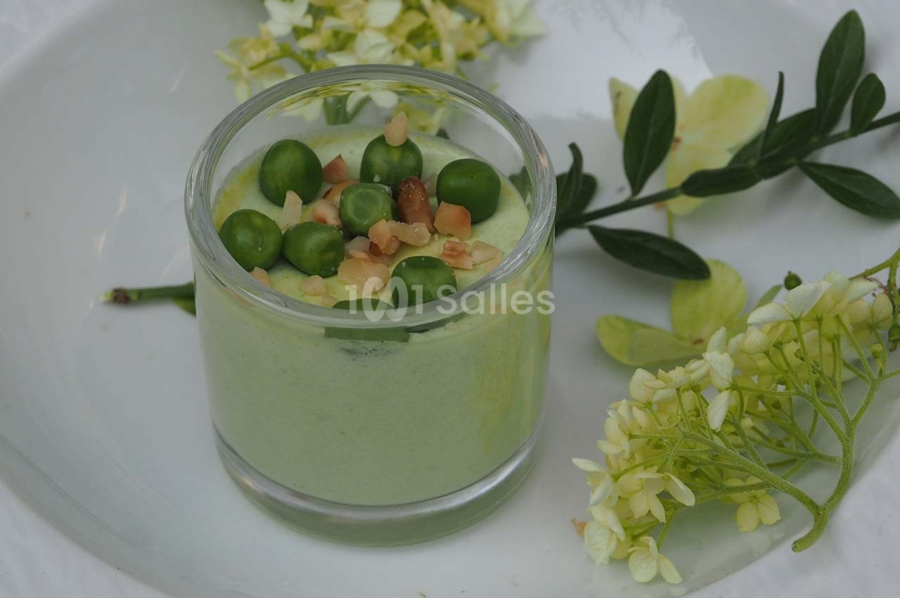 Verrine de mousse verte garnie de petits pois et éclats de noisettes, entourée de fleurs et feuilles décoratives.