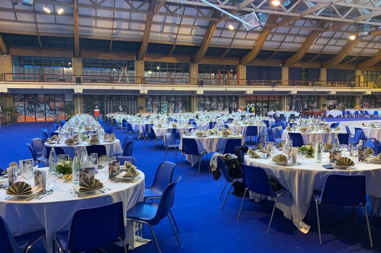 Salle de réception avec des tables rondes dressées, nappes blanches, chaises bleues et décorations élégantes.
