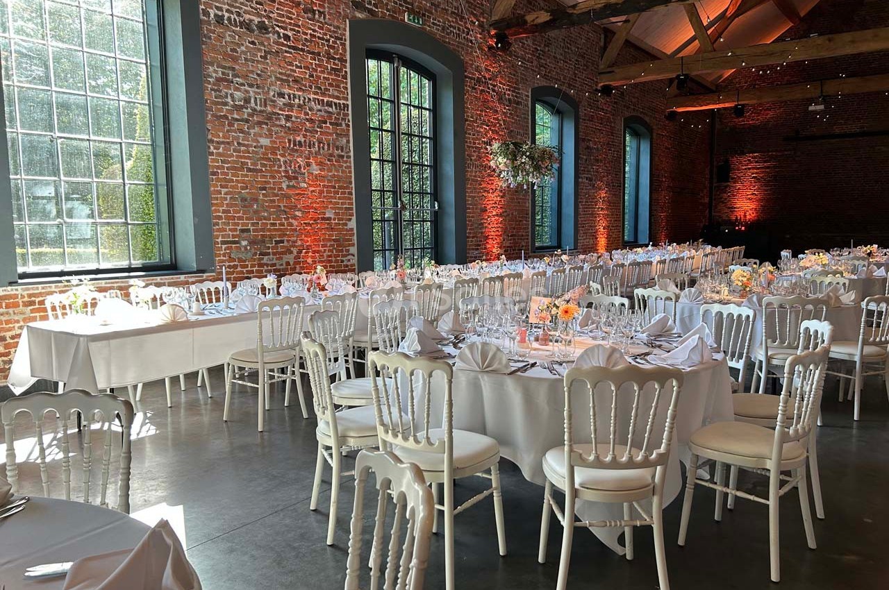 Salle de réception avec tables rondes dressées, chaises blanches et murs en briques sous un plafond en poutres apparentes.