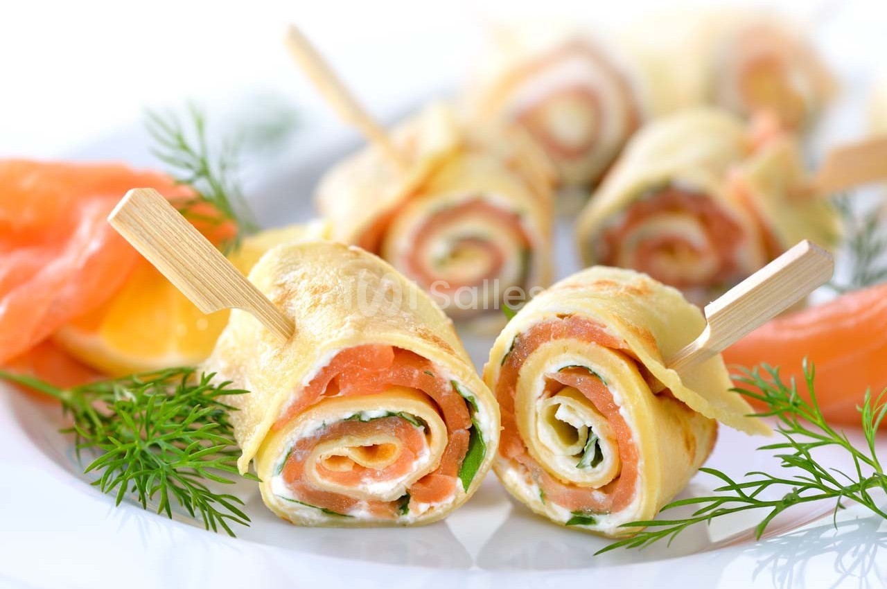 Roulés de crêpes garnis de saumon fumé et d'herbes, présentés sur un plateau avec des piques en bois.