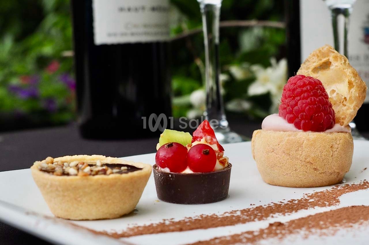 Assortiment de desserts miniatures sur une assiette blanche, décorée de cacao en poudre, avec un fond flou de bouteilles de…
