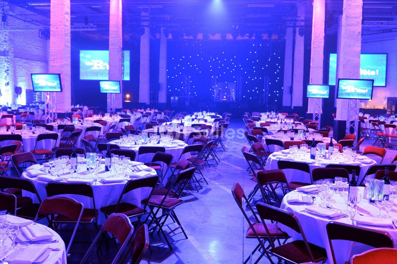 Salle de réception éclairée en violet, avec des tables rondes dressées et des écrans disposés autour.