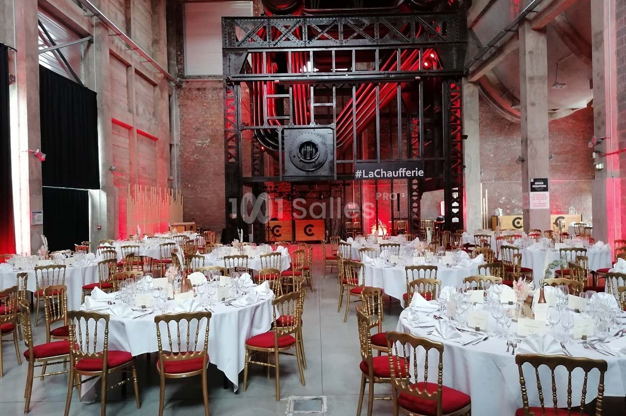 Salle industrielle aménagée pour un événement, avec des tables rondes dressées et des chaises dorées.