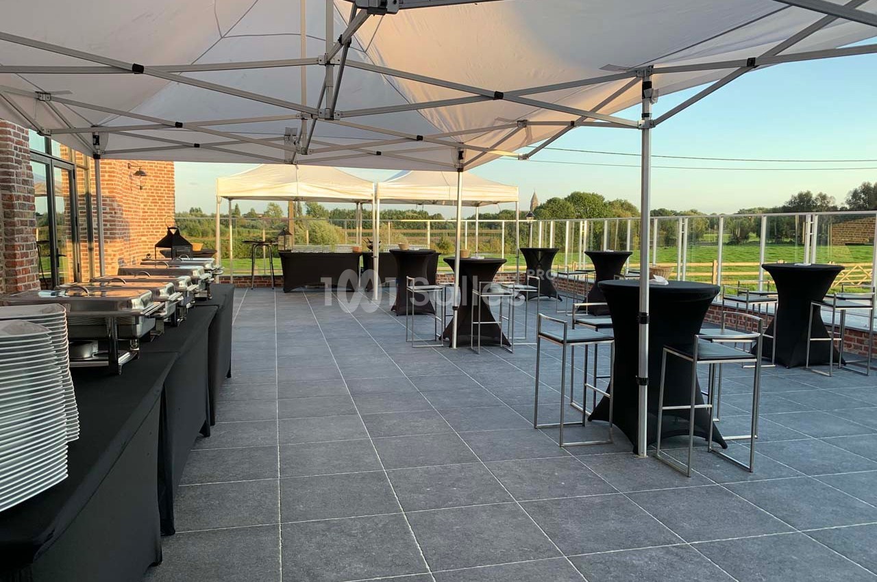 Terrasse aménagée avec des tables hautes, des buffets et des tentes blanches, offrant une vue sur un paysage verdoyant.