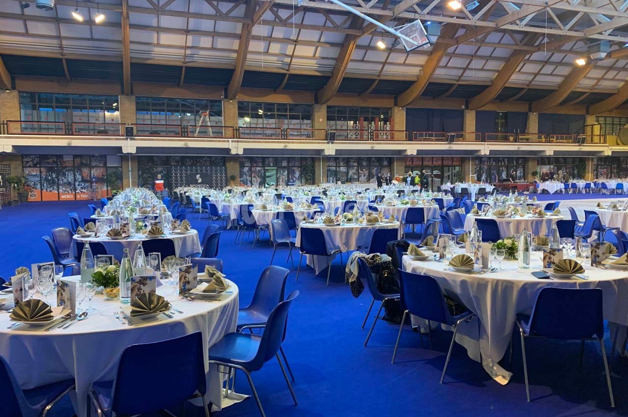 Salle de réception avec tables rondes dressées, nappes blanches, chaises bleues et décorations élégantes.