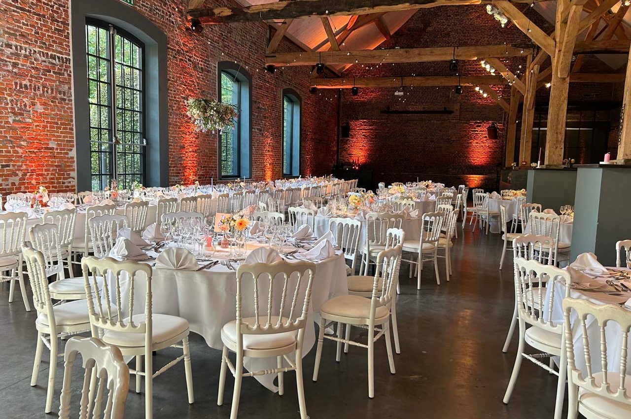 Salle de réception avec murs en briques, grandes fenêtres et tables rondes dressées pour un événement.
