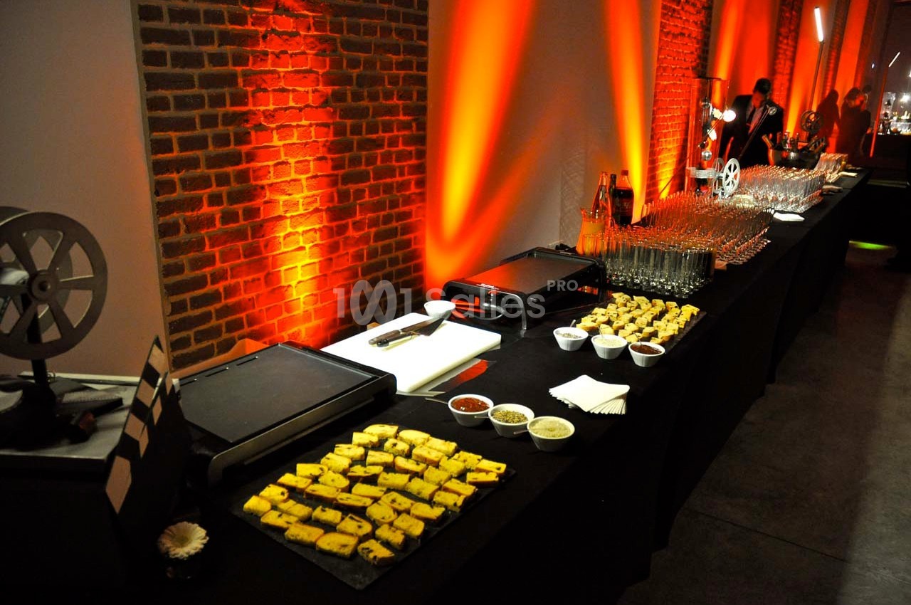 Buffet avec planchas, amuse-bouches, sauces et verres, disposé dans une salle éclairée par des lumières orange.