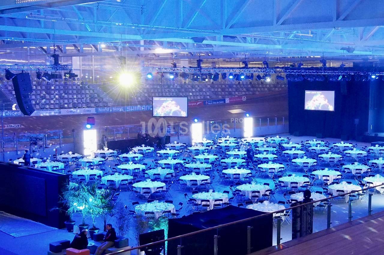 Salle de réception illuminée en bleu avec des tables rondes dressées, située dans un espace sportif couvert.